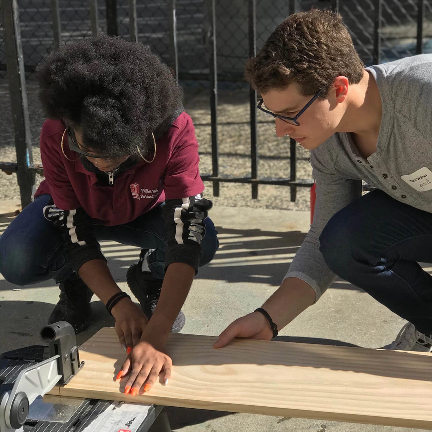 Two people crouch on a sunny sidewalk measuring a wooden plank on a saw stand, as a volunteer wearing a “Zach Wilner” name tag guides a young student in a maroon school polo.