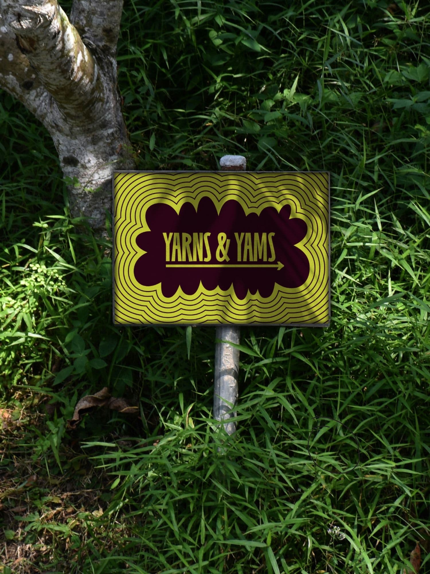 Outdoor garden sign reading YARNS & YAMS with a yellow and maroon wavy pattern, planted in grass beside a tree.