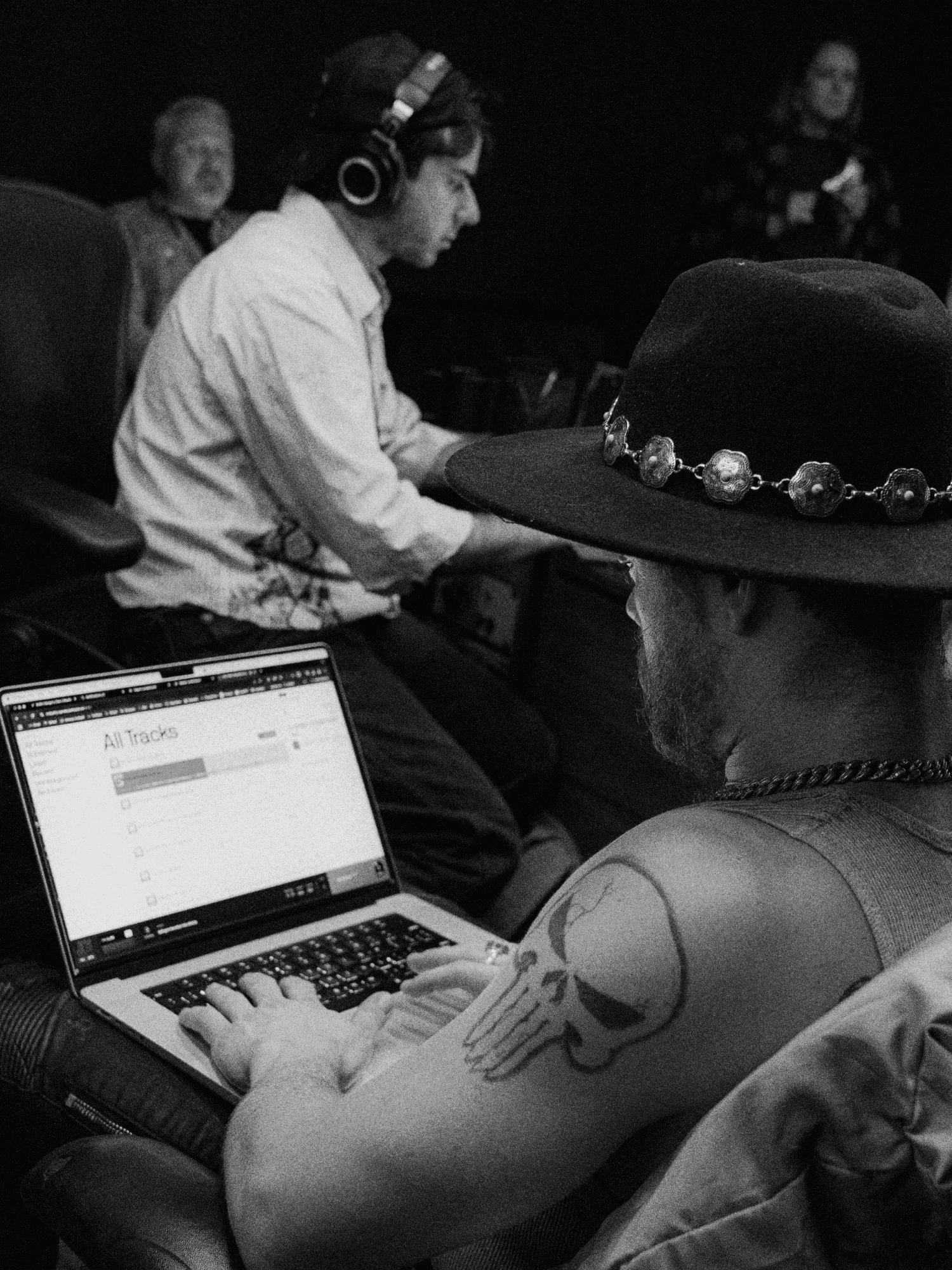 A black-and-white studio photo of a person in a wide-brim hat working at a laptop.