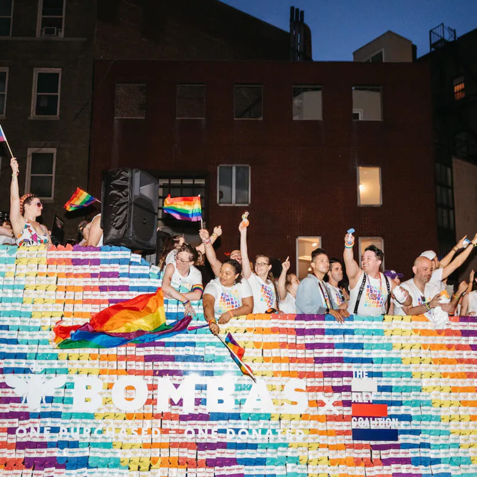 A nighttime Bombas Pride parade float decorated in rainbow rows of sock packaging, where people in white Bombas t-shirts wave rainbow flags beneath text reading “Bombas — One Purchased = One Donated” and “The Ally Coalition.”