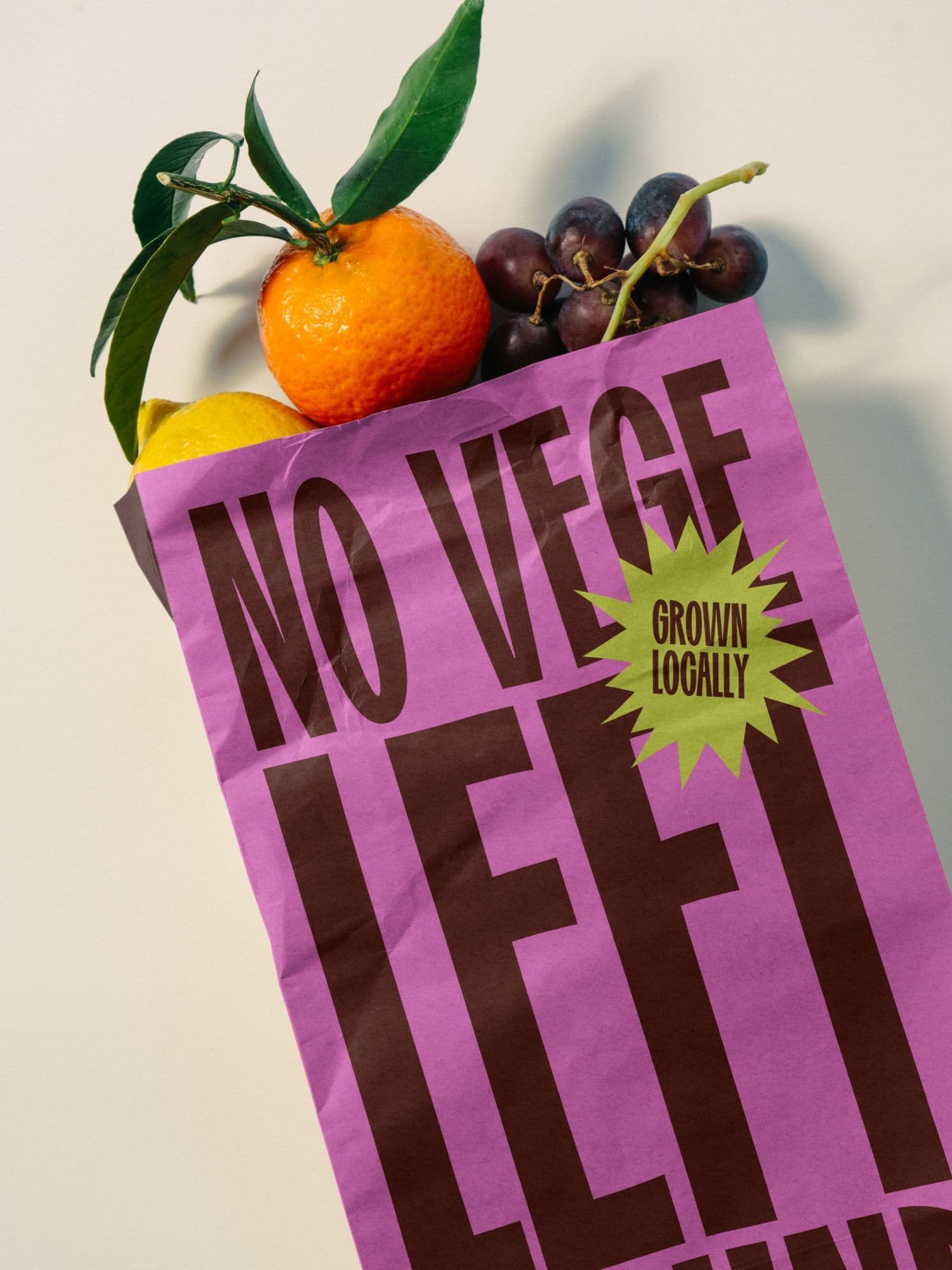 Bright pink paper bag printed with NO VEGE LEFT and a lime-green GROWN LOCALLY starburst, filled with an orange, lemon, and grapes.