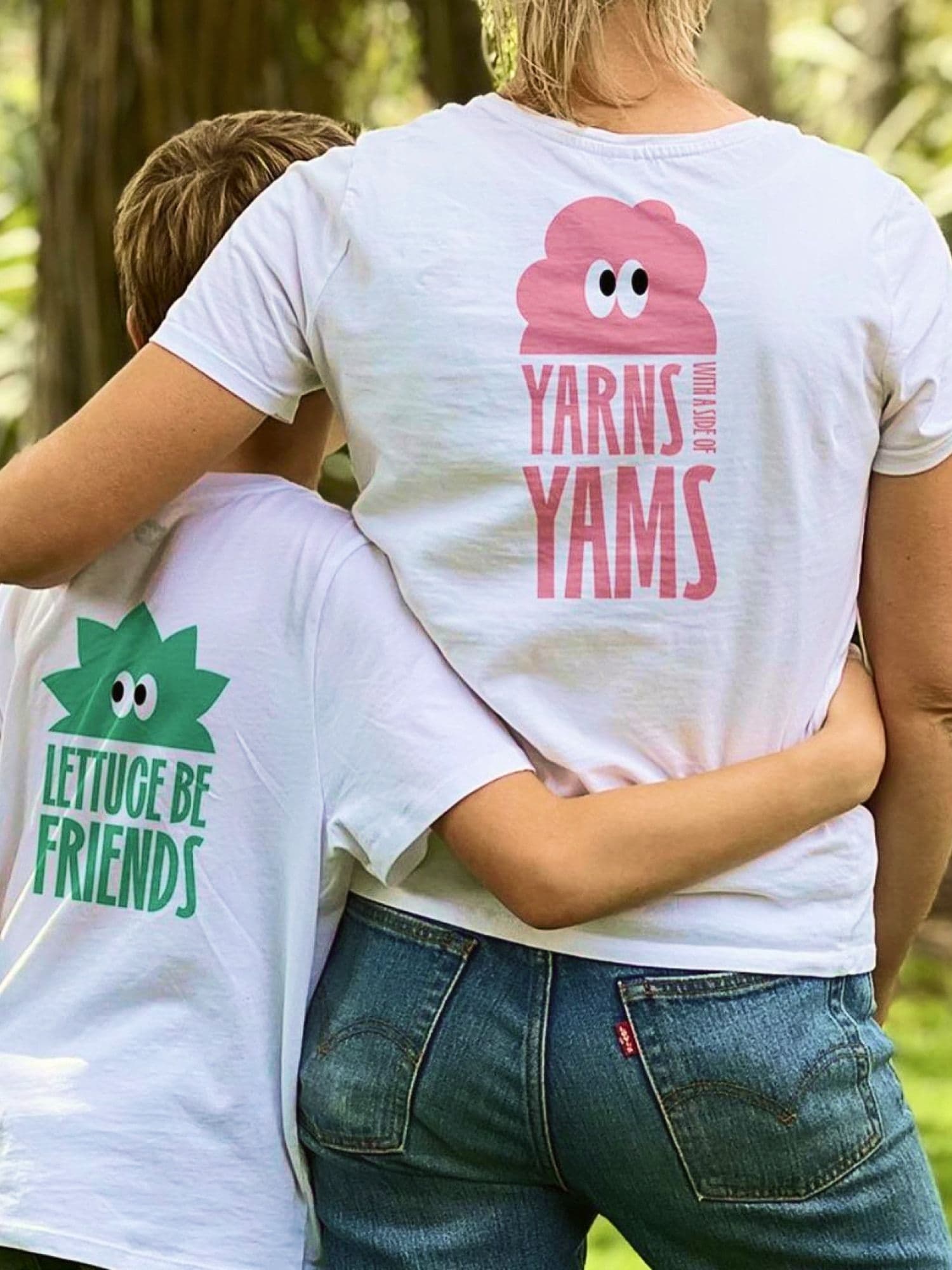 Woman and young boy seen from behind hugging in a garden, wearing white tees with lettuce and yam character puns.