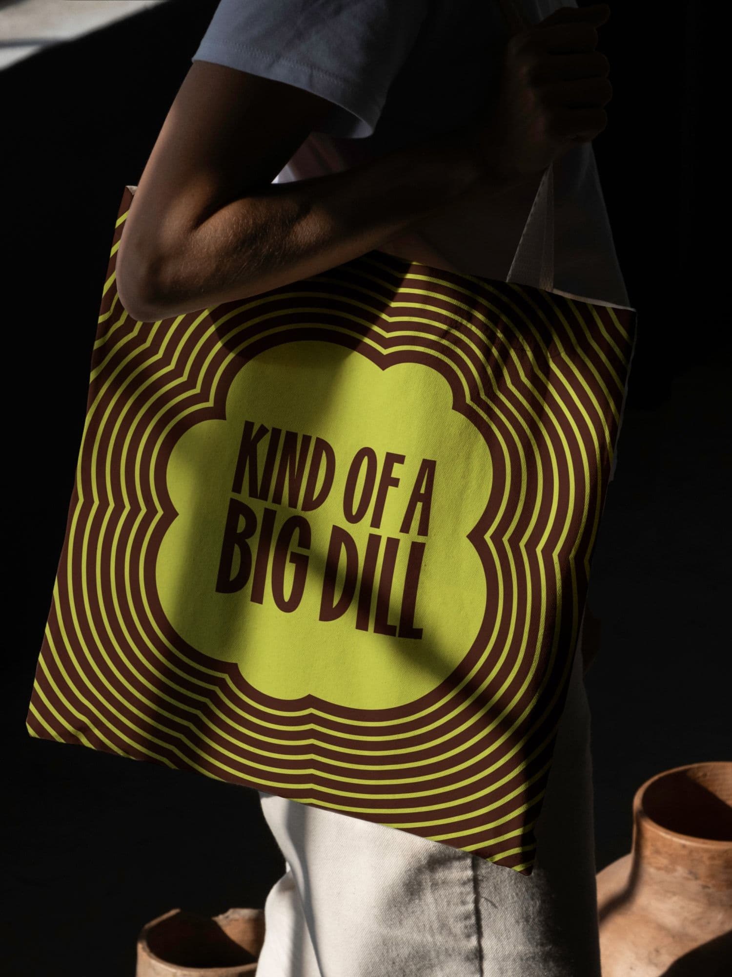 Person carrying a canvas tote with KIND OF A BIG DILL in a lime-green scalloped graphic surrounded by concentric brown and green waves.